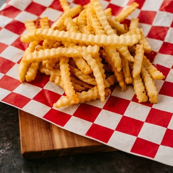 Crispy crinkle-cut french fries piled on red and white checkered paper, on a wooden board.