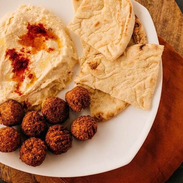 A white plate with hummus, falafel, and pita bread on a wooden board with an orange napkin.